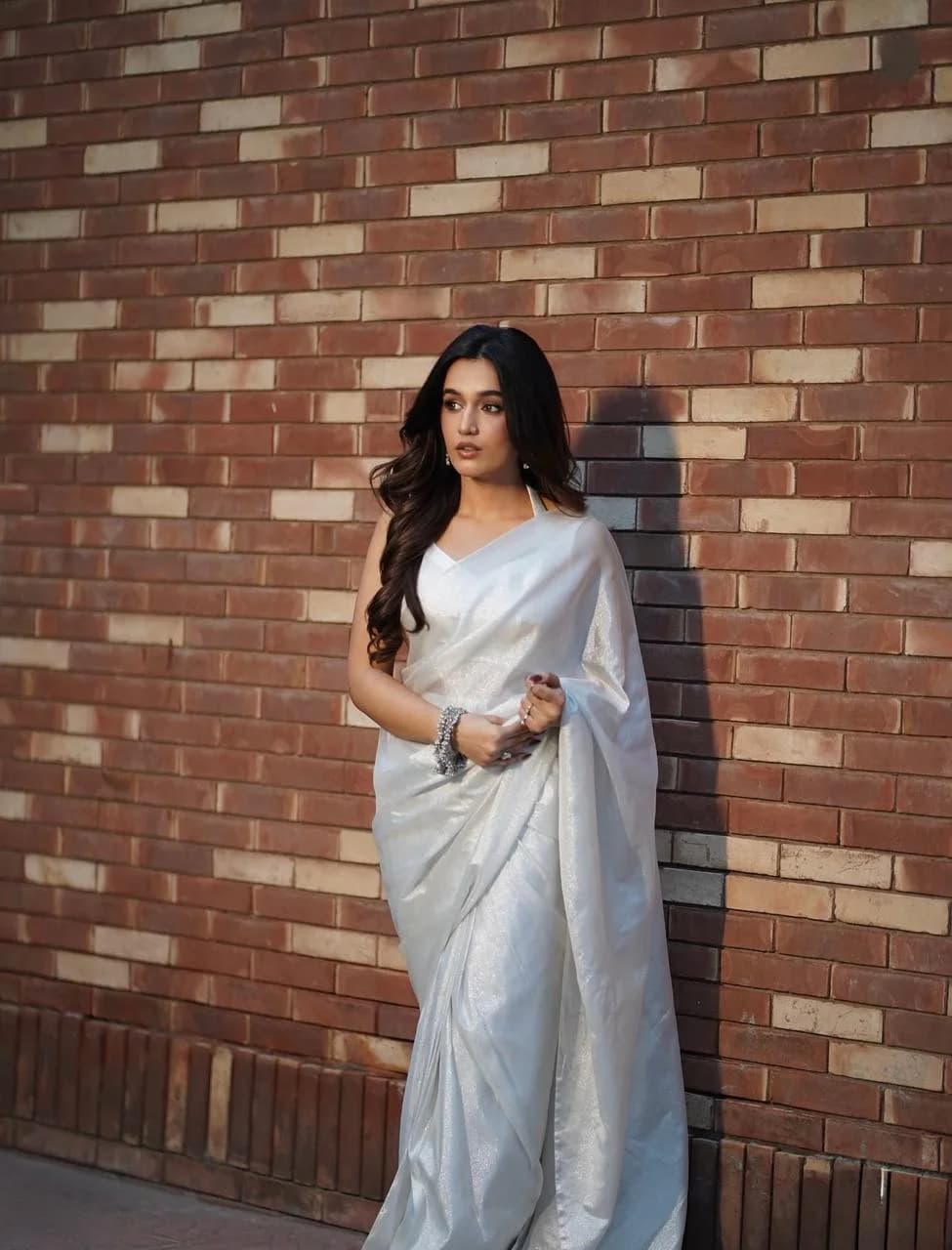 Woman in a shimmering silver saree standing against a rustic brick wall