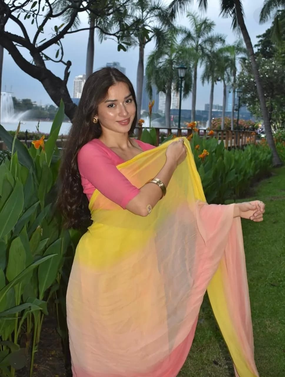 Woman in a pink and yellow ombré saree twirling in a garden with palm trees and a fountain